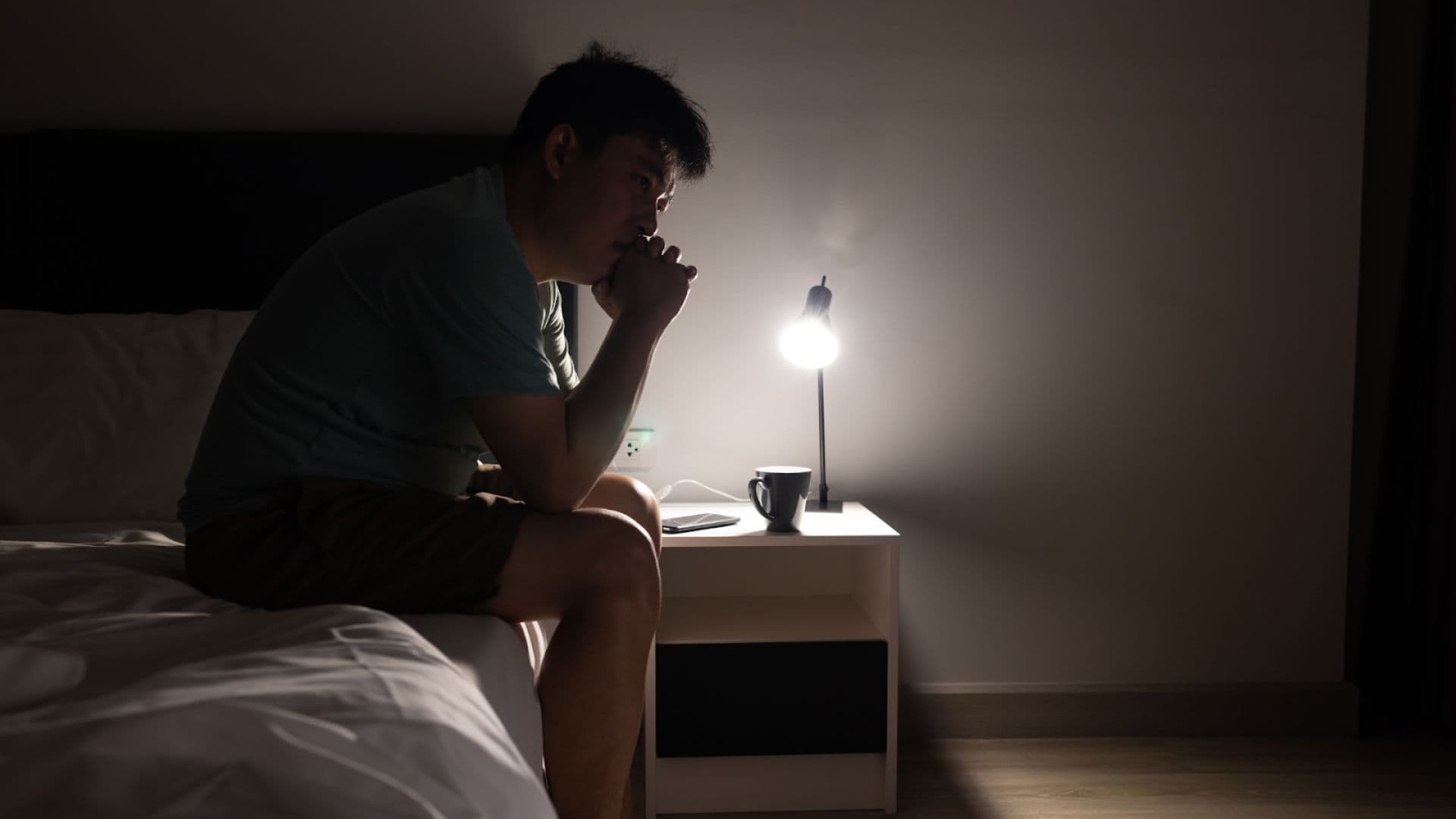 Seorang pria duduk di pinggir kasur sambil merenung di kamar yang gelap